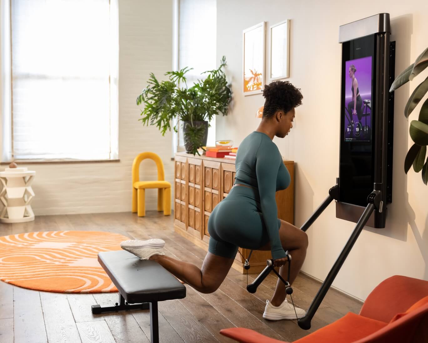 A woman is working out in her living room. She is facing Tonal and is completing a Bulgarian Split Squat.