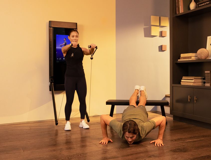 A couple works out in their living room. The woman is stands with her back to Tonal, completing a Front Raise. Beside her, the man is completing an Elevated Push Up using the Tonal bench.