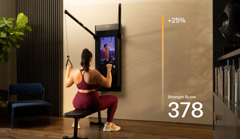 A woman is working out in her living room. She is sitting on the Tonal bench facing the machine and using the handles to complete a Lat Pulldown.